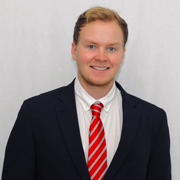 Headshot of Owen Gleeson dressed in a red tie and black suit against a gray background.